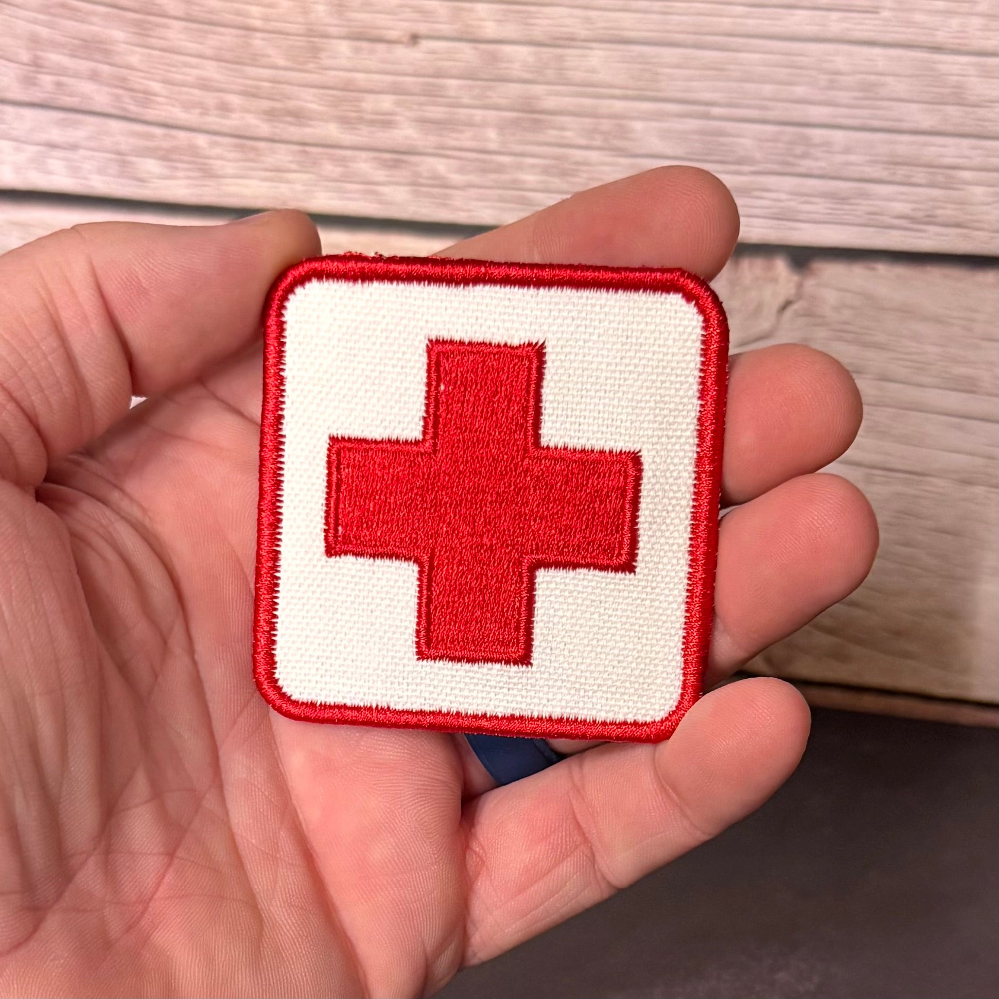 A square embroidered patch with a red cross on a white background, held in a person's hand against a light wood surface.