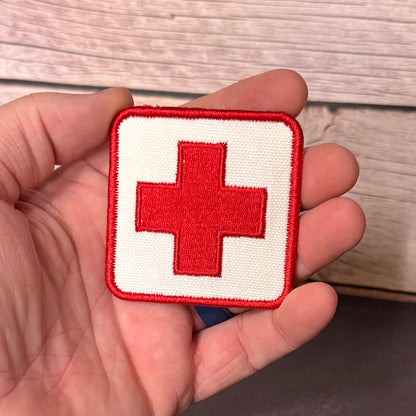 A square embroidered patch with a red cross on a white background, held in a person's hand against a light wood surface.