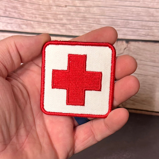 A square embroidered patch with a red cross on a white background, held in a person's hand against a light wood surface.