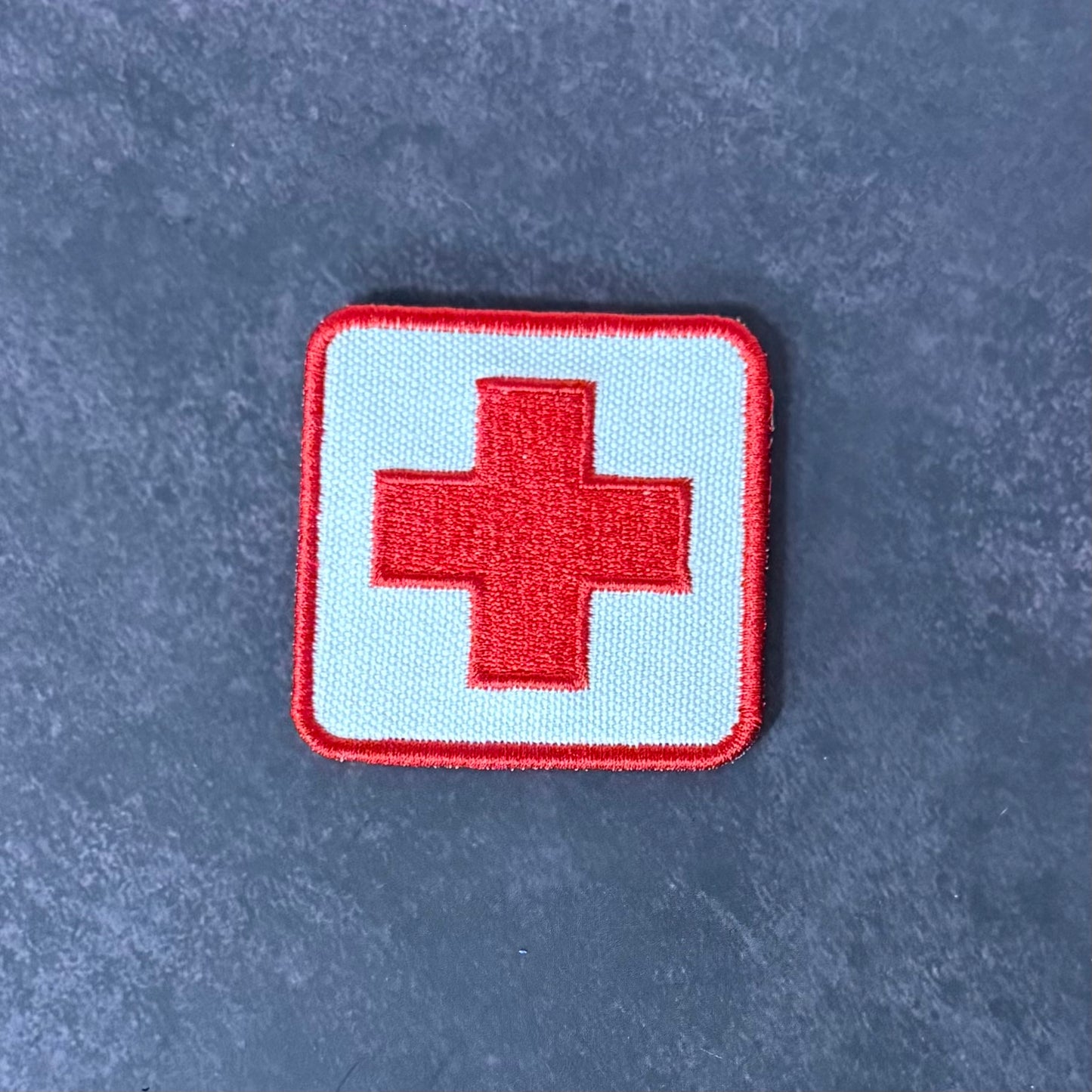 An embroidered square patch featuring a red cross on a light blue background with a red border, placed on a textured dark gray surface.