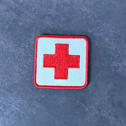 An embroidered square patch featuring a red cross on a light blue background with a red border, placed on a textured dark gray surface.