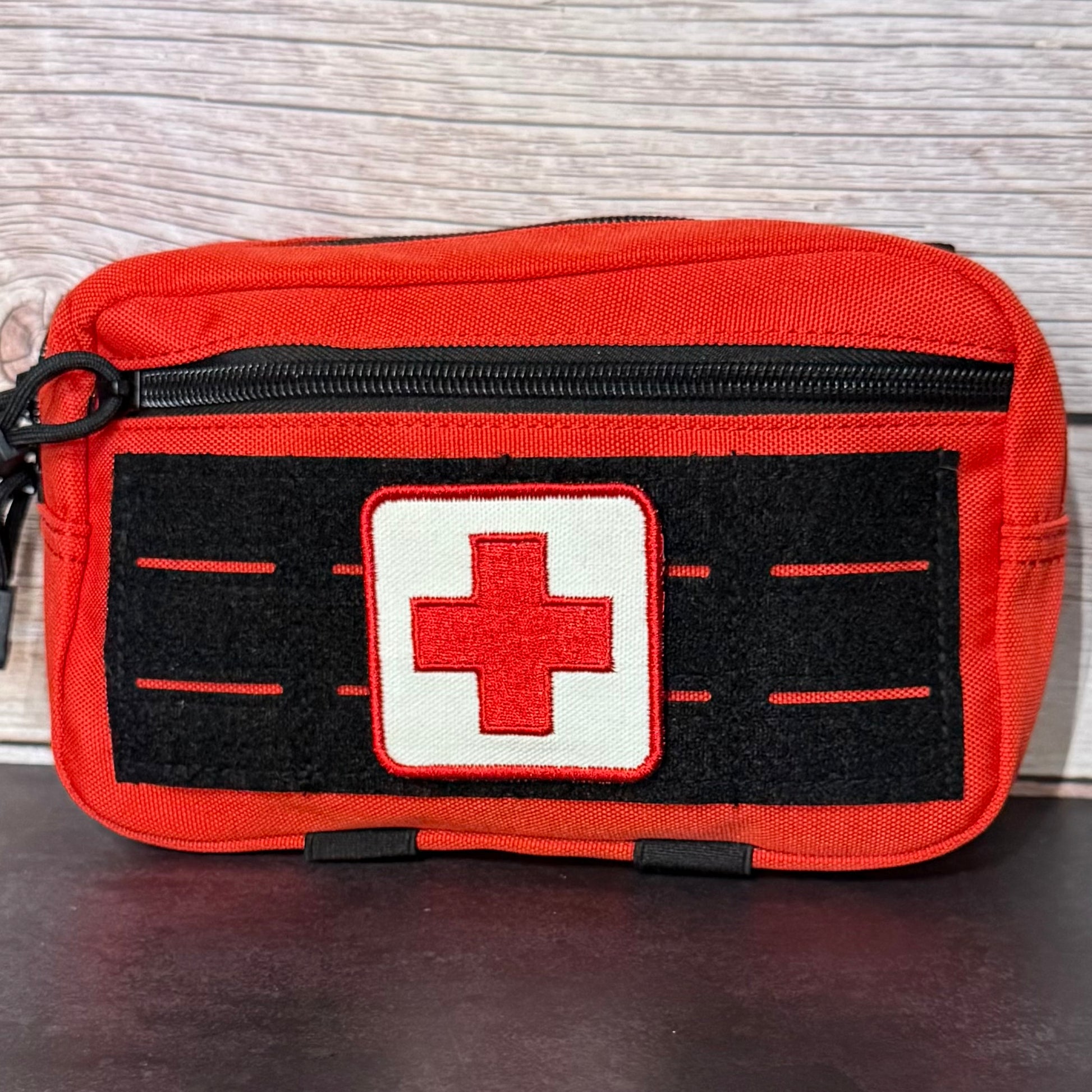 An orange-red first aid pouch with a black zipper, a black velcro panel, and a white square patch featuring a red cross in the center, shown against a light wood background.