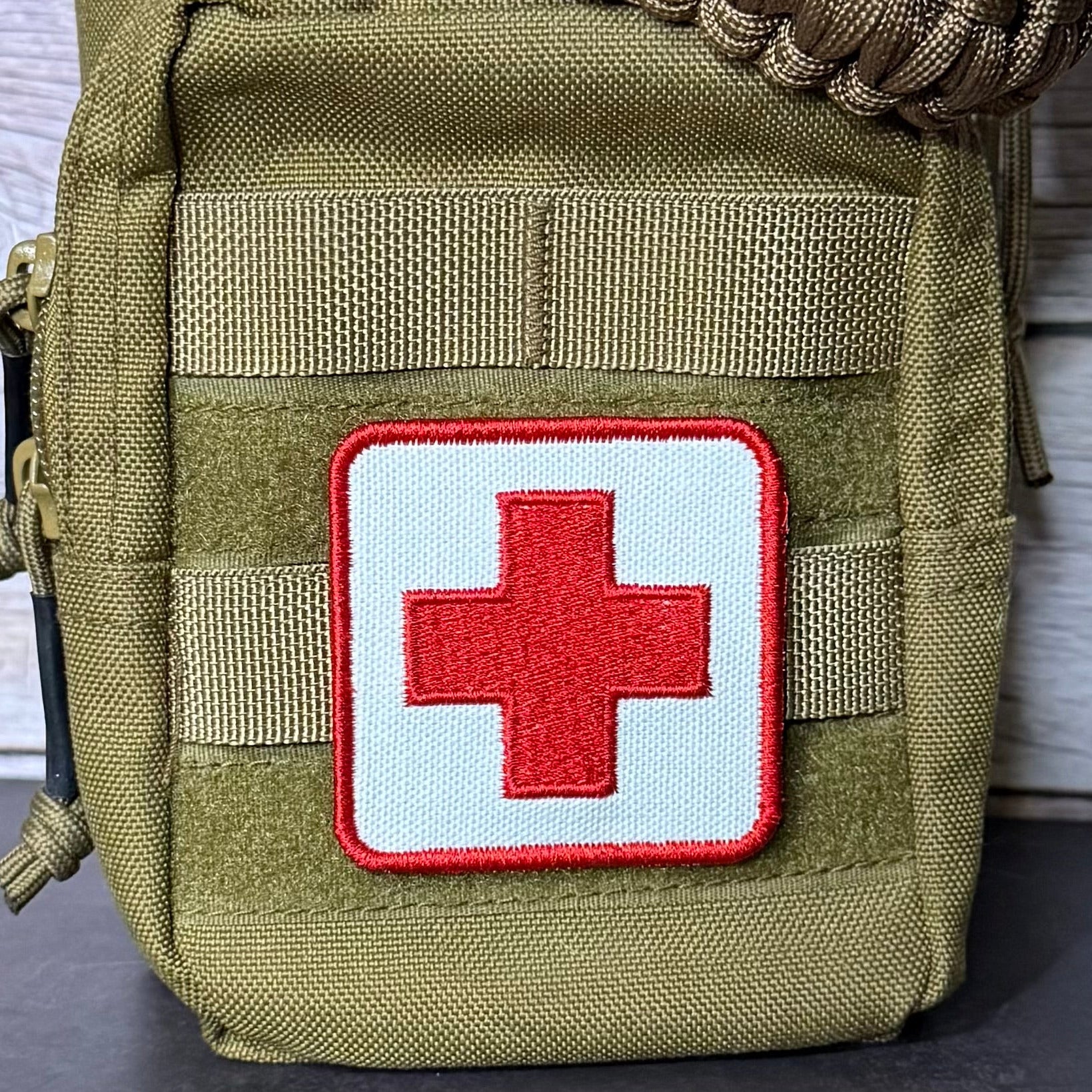 An olive green tactical pouch with a white square patch featuring a red cross, set against a light wood background.