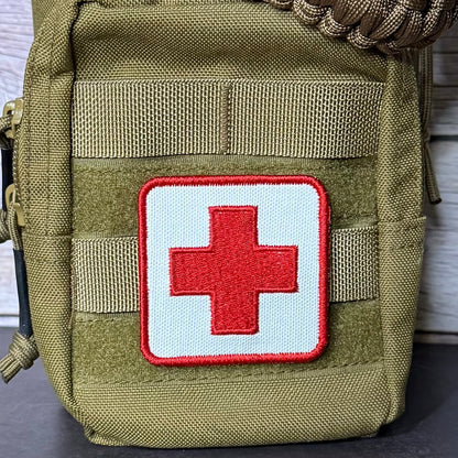An olive green tactical pouch with a white square patch featuring a red cross, set against a light wood background.