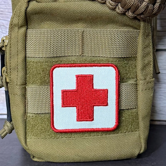 An olive green tactical pouch with a white square patch featuring a red cross, set against a light wood background.