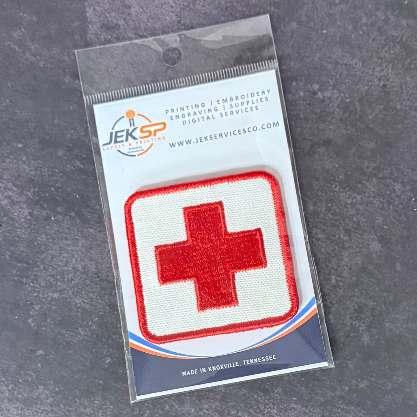 A red and white embroidered medical cross patch in its original clear plastic retail packaging, displayed on a dark gray textured surface.