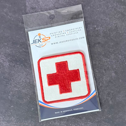 A red and white embroidered medical cross patch in its original clear plastic retail packaging, displayed on a dark gray textured surface.