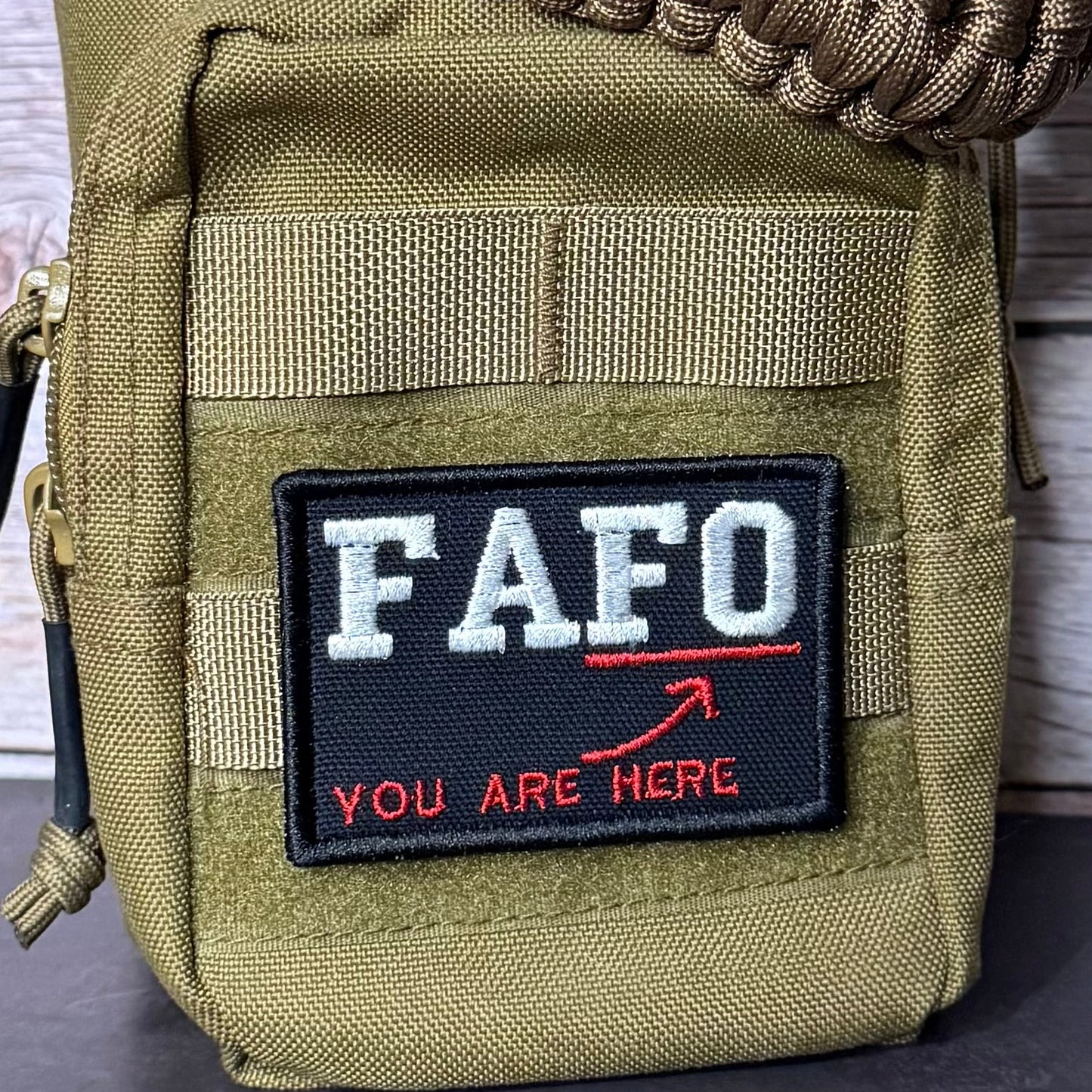A close-up of a tactical utility pouch in olive drab green nylon, featuring a black rectangular patch with the white text 'FAFO' and red text 'YOU ARE HERE' with a red arrow. A brown paracord strap is wrapped around the top, and a zipper pull is visible on the left side.