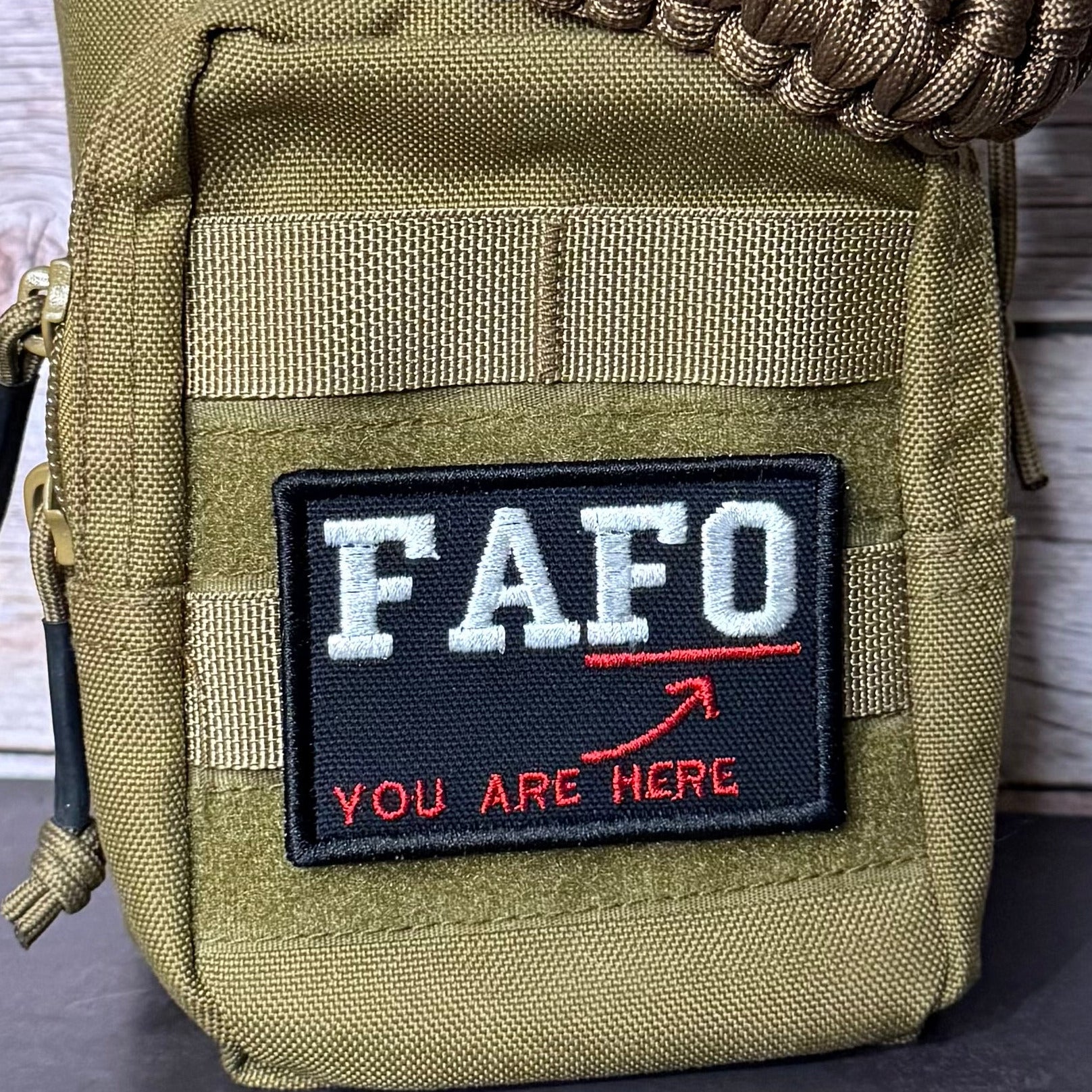 A close-up of a tactical utility pouch in olive drab green nylon, featuring a black rectangular patch with the white text 'FAFO' and red text 'YOU ARE HERE' with a red arrow. A brown paracord strap is wrapped around the top, and a zipper pull is visible on the left side.