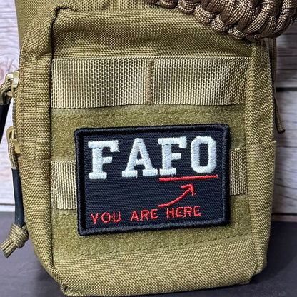 A close-up of a tactical utility pouch in olive drab green nylon, featuring a black rectangular patch with the white text 'FAFO' and red text 'YOU ARE HERE' with a red arrow. A brown paracord strap is wrapped around the top, and a zipper pull is visible on the left side.