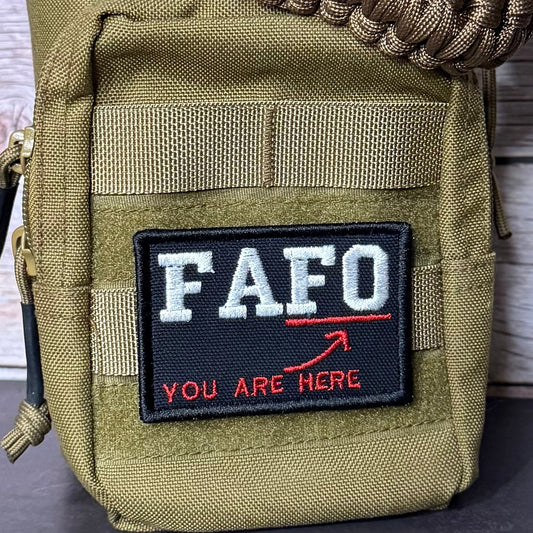 A close-up of a tactical utility pouch in olive drab green nylon, featuring a black rectangular patch with the white text 'FAFO' and red text 'YOU ARE HERE' with a red arrow. A brown paracord strap is wrapped around the top, and a zipper pull is visible on the left side.
