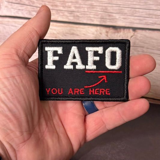 A hand holding a rectangular black embroidered patch with white 'FAFO' lettering, a red underline, and red text reading 'YOU ARE HERE' with a red arrow, against a light wood background.