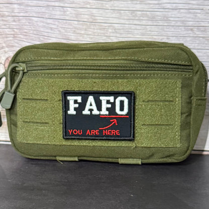 An olive green tactical pouch with a black rectangular patch featuring the white text 'FAFO' and red text 'YOU ARE HERE' with a red arrow, set against a light wood background.