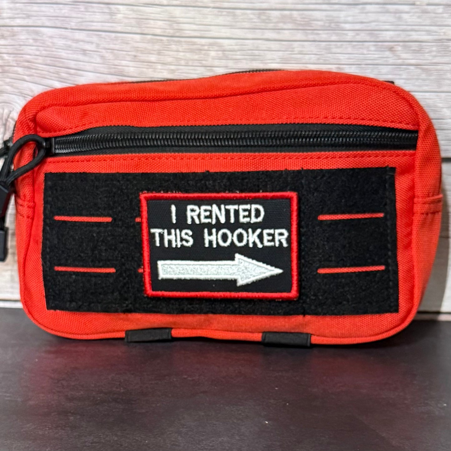 A red zippered pouch with a black velcro patch that reads 'I RENTED THIS HOOKER' with a white arrow, placed on a dark surface against a light wood background.