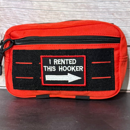 A red zippered pouch with a black velcro patch that reads 'I RENTED THIS HOOKER' with a white arrow, placed on a dark surface against a light wood background.