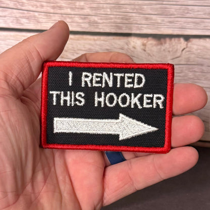 A hand holding a rectangular embroidered patch with a black background, red border, white text reading 'I RENTED THIS HOOKER,' and a white arrow pointing right, against a wood plank background.