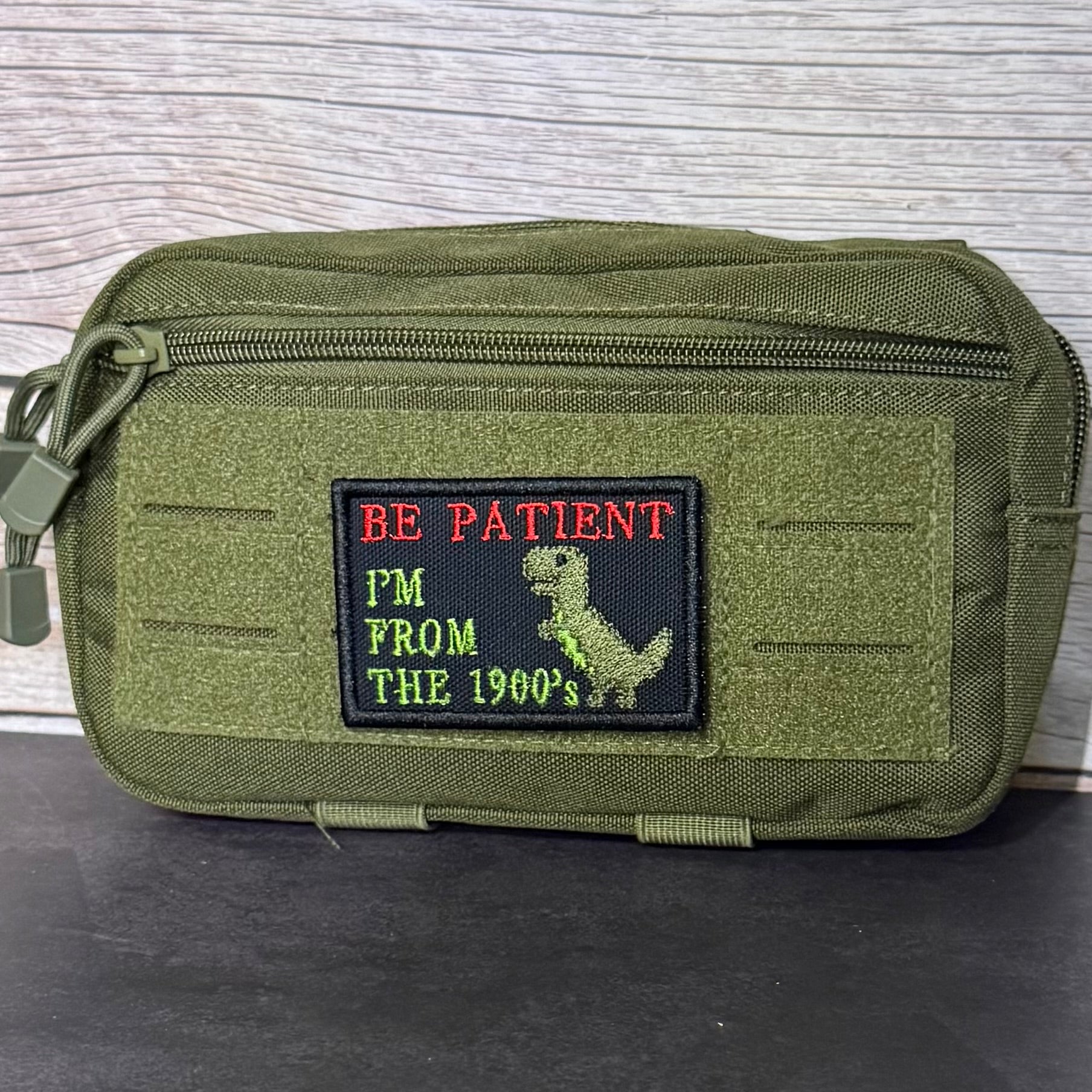 An olive green tactical pouch with a front zipper and a rectangular embroidered patch that reads 'BE PATIENT I'M FROM THE 1900s' with a dinosaur graphic, placed on a dark surface against a light wood background.