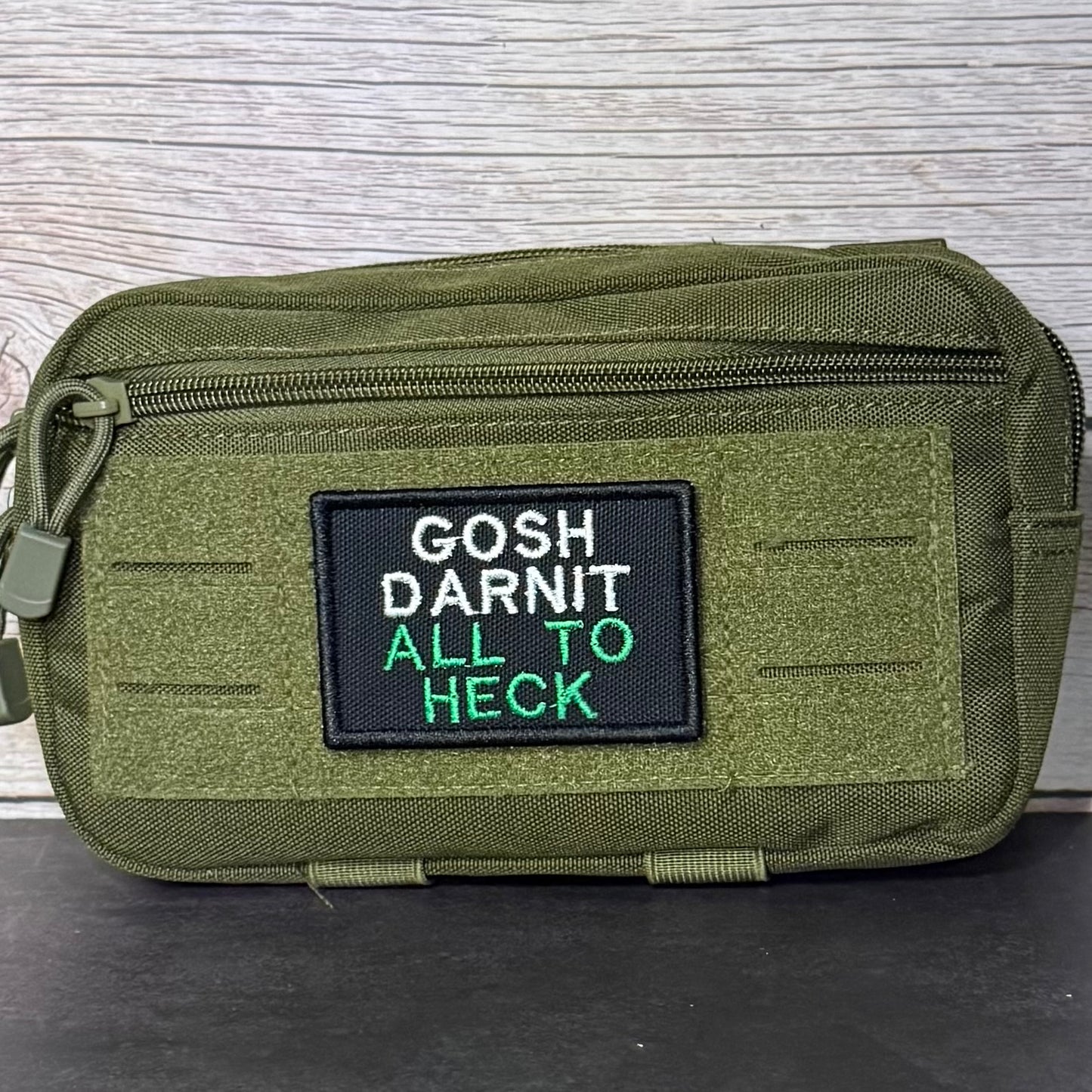 An olive green tactical pouch with a black rectangular patch featuring the embroidered text 'GOSH DARNIT ALL TO HECK' in white and green, set against a light wood background.