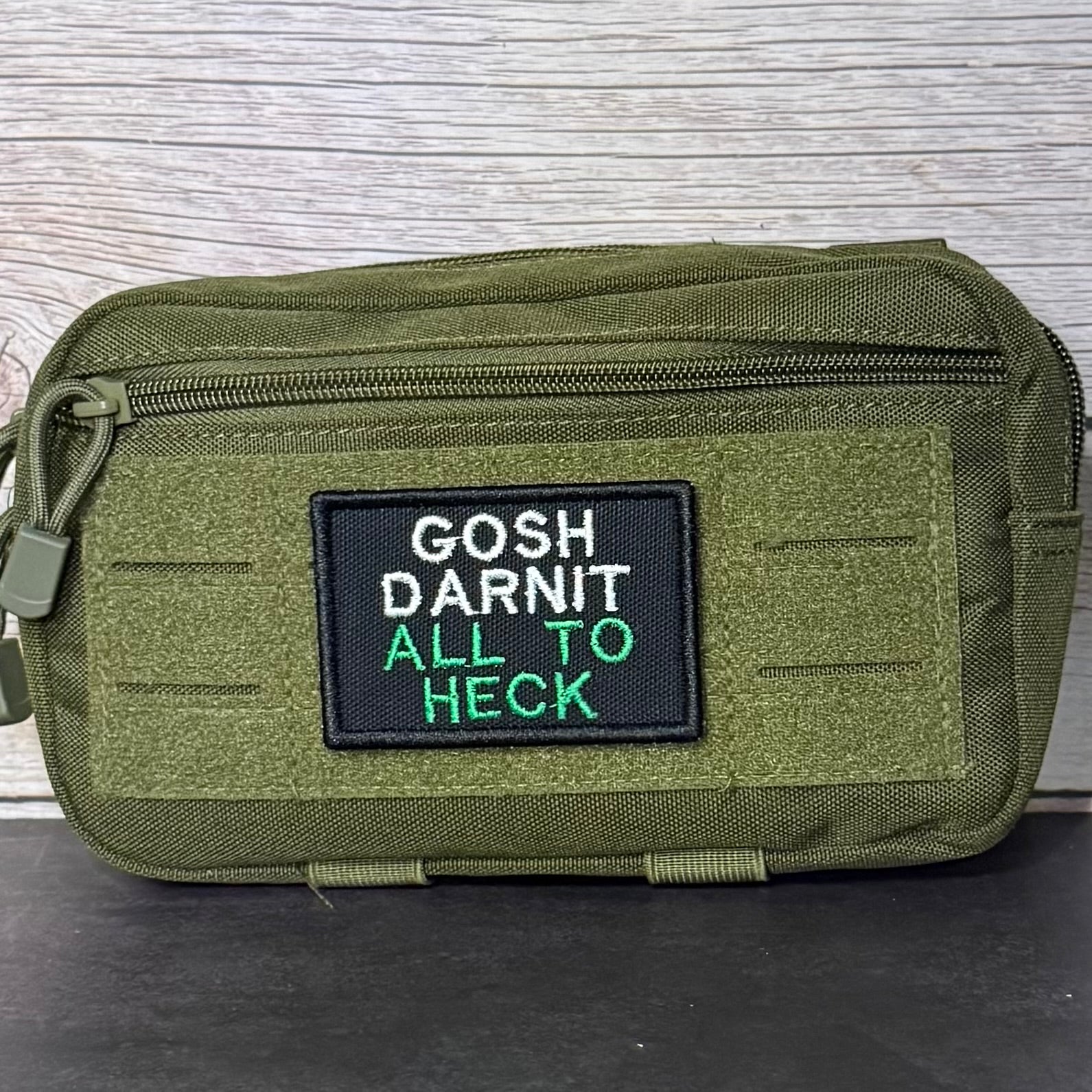 An olive green tactical pouch with a black rectangular patch featuring the embroidered text 'GOSH DARNIT ALL TO HECK' in white and green, set against a light wood background.