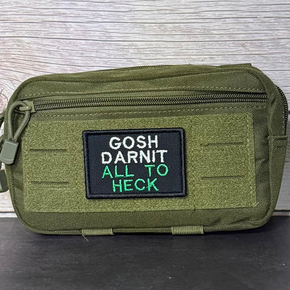 An olive green tactical pouch with a black rectangular patch featuring the embroidered text 'GOSH DARNIT ALL TO HECK' in white and green, set against a light wood background.