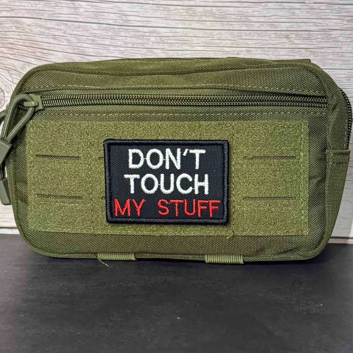 An olive green tactical pouch with a black embroidered patch that reads 'DON'T TOUCH MY STUFF' in white and red text, set against a gray wood-grain background.