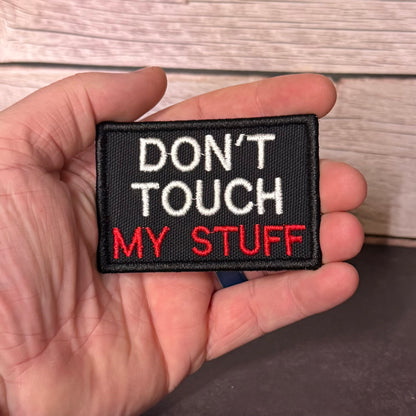 A hand holding a rectangular embroidered patch with the text 'DON'T TOUCH MY STUFF' in white and red on a black background, with a light wood plank surface behind it.