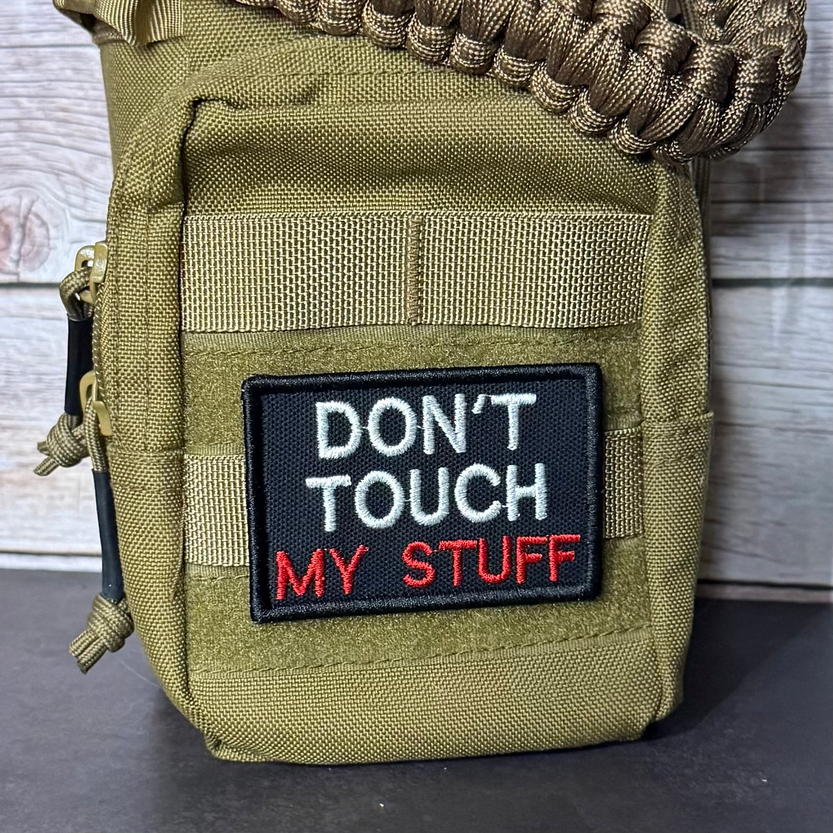 An olive green tactical pouch with a black embroidered patch that reads 'DON'T TOUCH MY STUFF' in white and red text. A brown paracord strap is threaded through the top zipper, and the pouch is photographed against a rustic wood plank background.