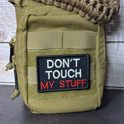An olive green tactical pouch with a black embroidered patch that reads 'DON'T TOUCH MY STUFF' in white and red text. A brown paracord strap is threaded through the top zipper, and the pouch is photographed against a rustic wood plank background.