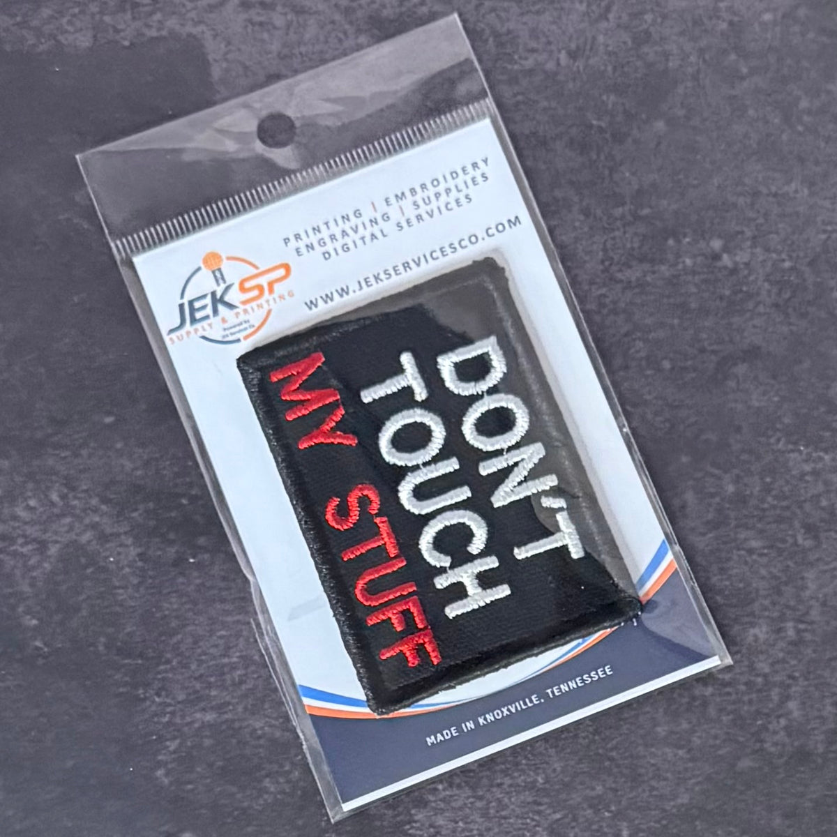 A black rectangular embroidered patch with the phrase 'MY TOUCH DON'T PUNCH' in white and red lettering, sealed in a clear plastic retail package with a branded card insert on a dark textured surface.