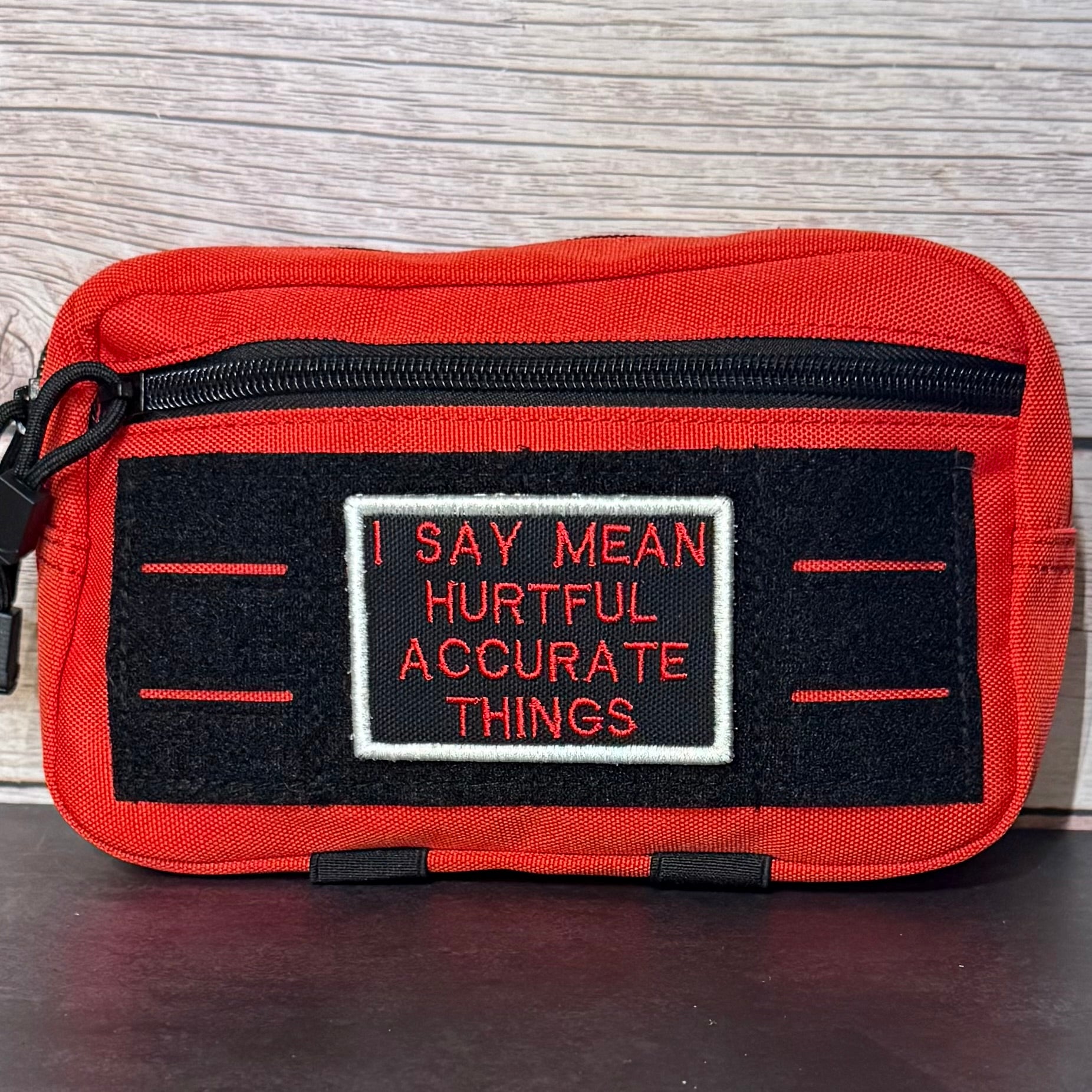 A red zippered pouch with a black velcro patch that reads 'I SAY MEAN HURTFUL ACCURATE THINGS' in red and white embroidery, set against a wood-grain background.