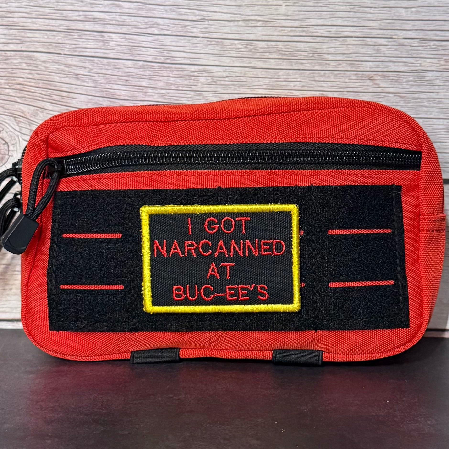 A red and black pouch featuring a novelty embroidered patch that reads 'I GOT NARCANNED AT BUC-EE'S' in red text with a yellow border, set against a light wood background.