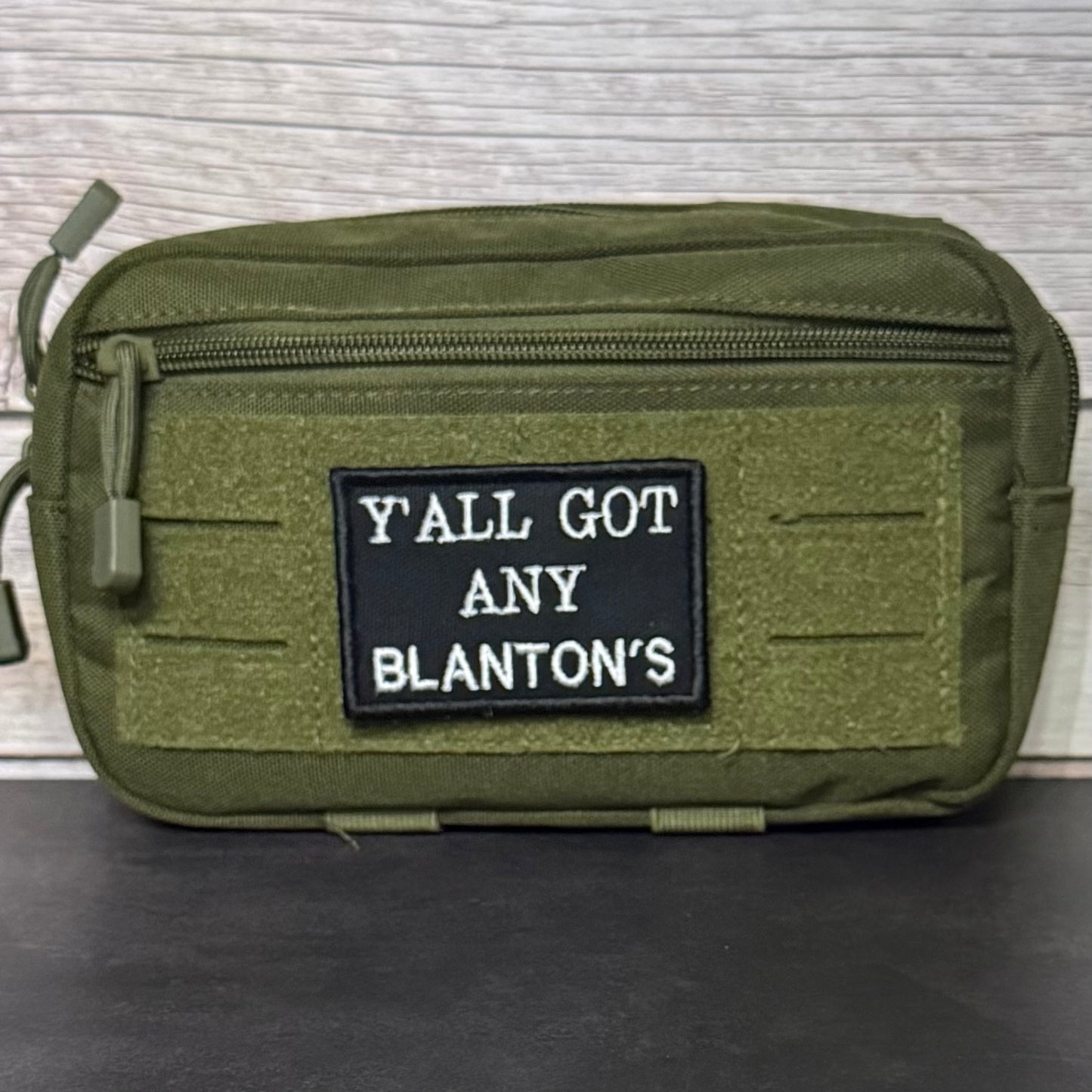 An olive green tactical pouch with a black rectangular patch featuring the white text 'Y'ALL GOT ANY BLANTON'S', displayed on a dark surface against a light wood background.