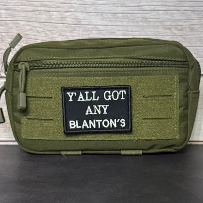An olive green tactical pouch with a black rectangular patch featuring the white text 'Y'ALL GOT ANY BLANTON'S', displayed on a dark surface against a light wood background.
