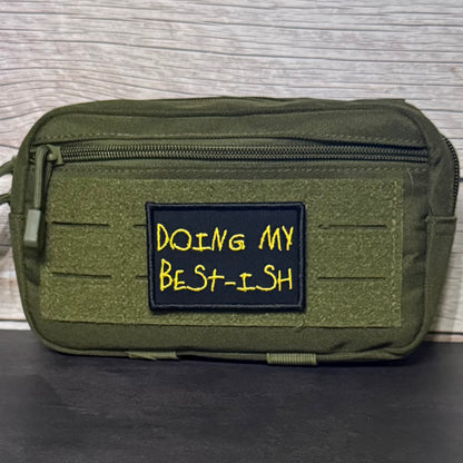 An olive green tactical pouch with a front zipper and a black rectangular patch embroidered with yellow text reading 'DOING MY BEST-ISH' against a wood-grain background.