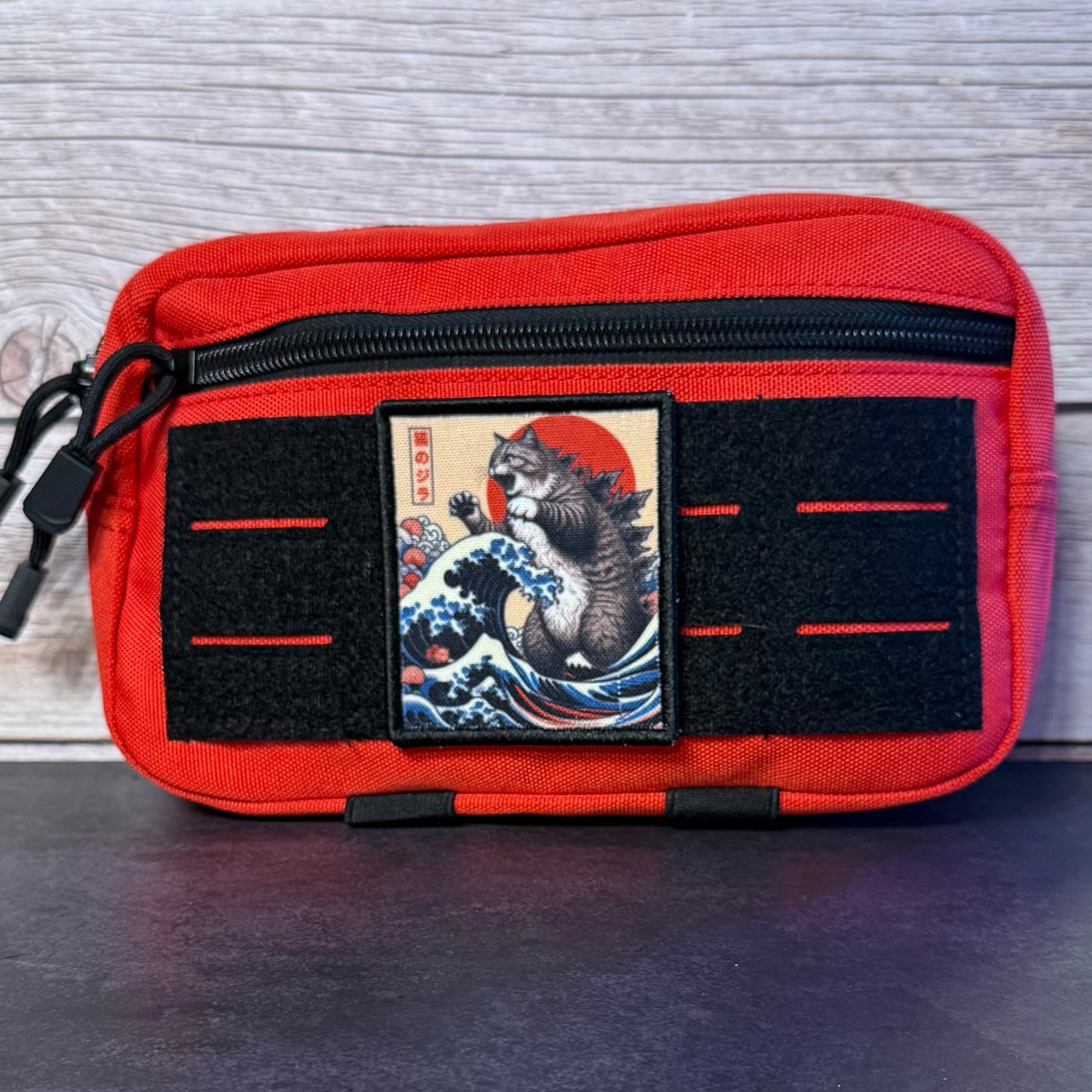 A red canvas utility pouch with black zippers and velcro strips, featuring a central patch with a Japanese-style cat illustration on a wave and red sun.