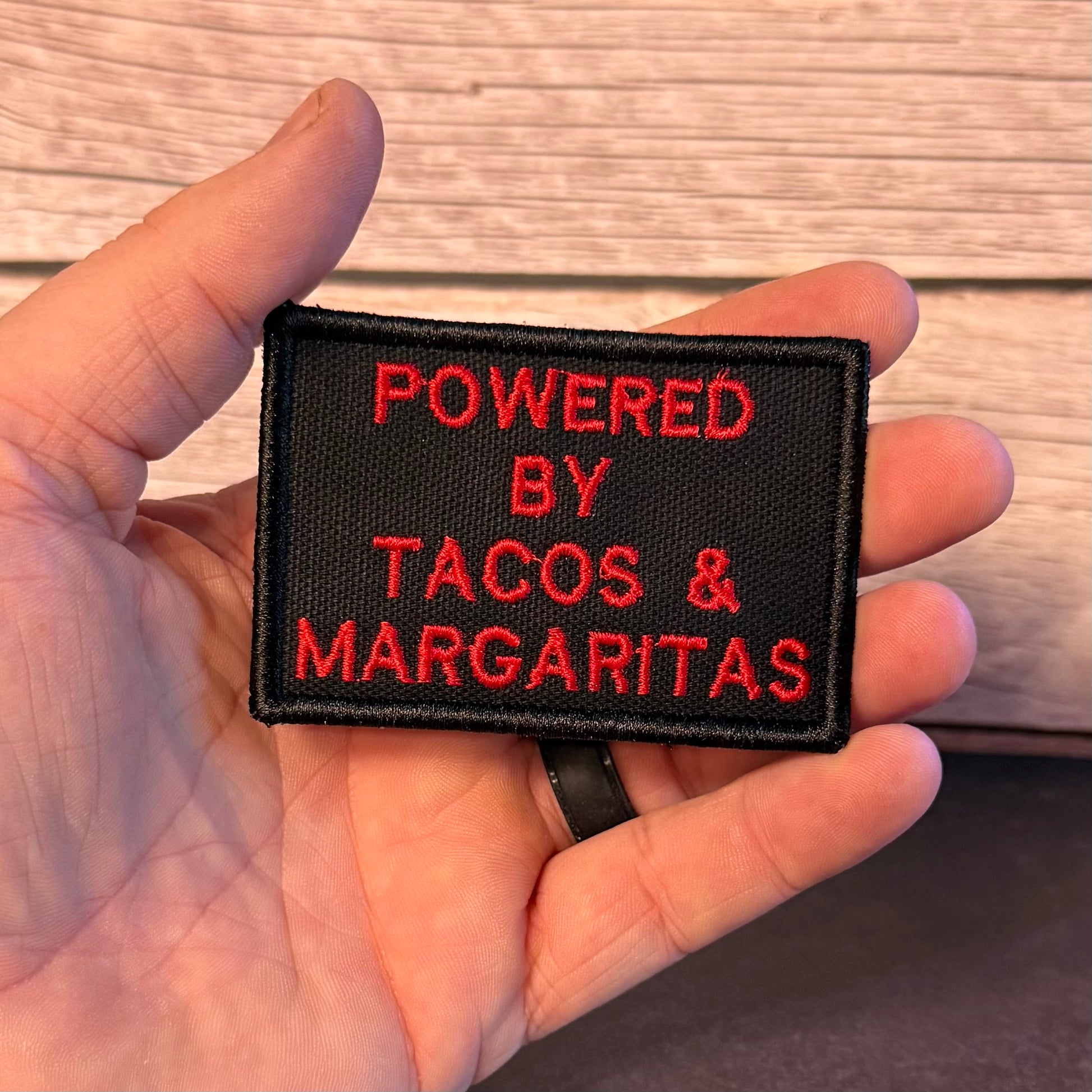 A hand holding a rectangular black embroidered patch with red text that reads 'POWERED BY TACOS & MARGARITAS' against a light wood background.