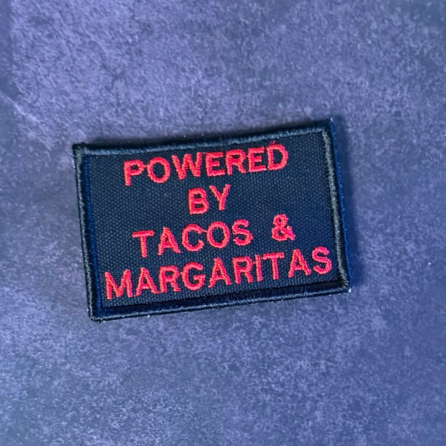 A rectangular embroidered patch with a black background and red text reading 'POWERED BY TACOS & MARGARITAS' on a textured blue surface.