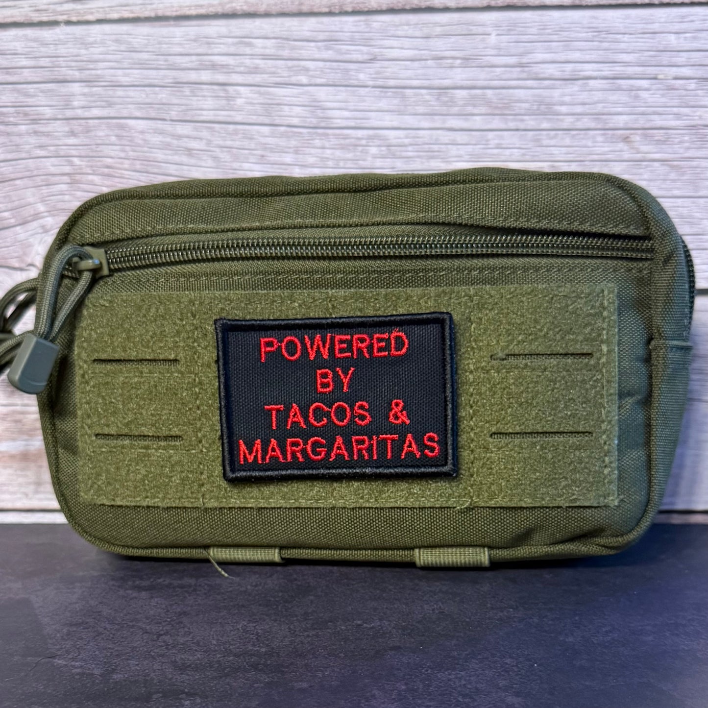 An olive green tactical pouch with a black rectangular patch featuring red embroidered text that reads 'POWERED BY TACOS & MARGARITAS', set against a light gray wood plank background.