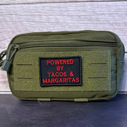 An olive green tactical pouch with a black rectangular patch featuring red embroidered text that reads 'POWERED BY TACOS & MARGARITAS', set against a light gray wood plank background.
