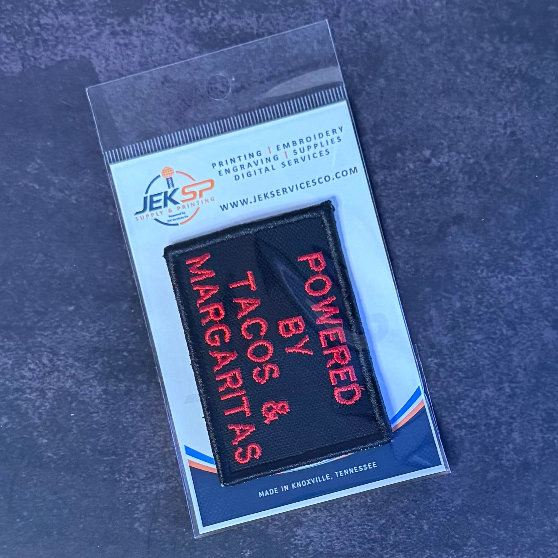 A black rectangular embroidered patch with red text reading 'POWERED BY TACOS & MARGARITAS' inside a clear plastic sleeve on a branded white backing card, photographed on a dark textured surface.