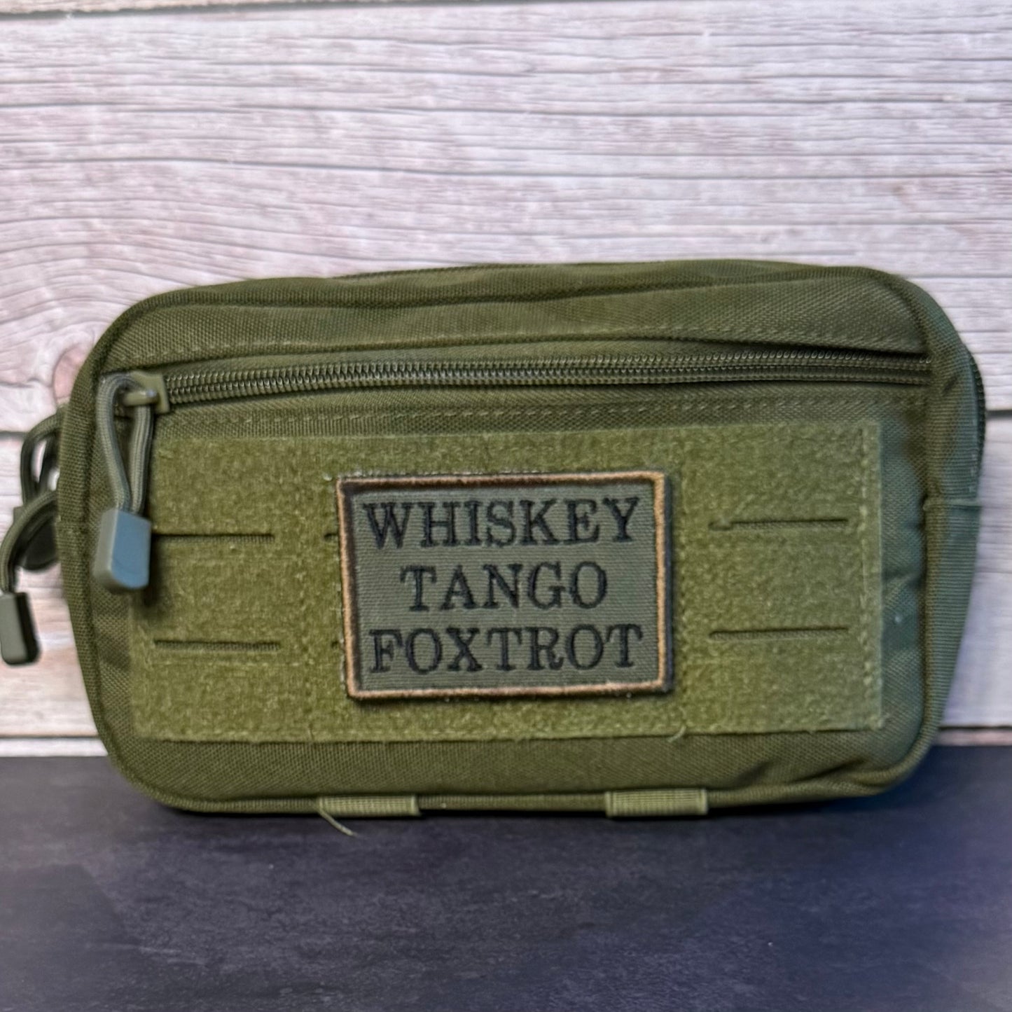 An olive green tactical pouch with a front zipper and a rectangular patch reading 'WHISKEY TANGO FOXTROT' in black letters, set against a light wood background.