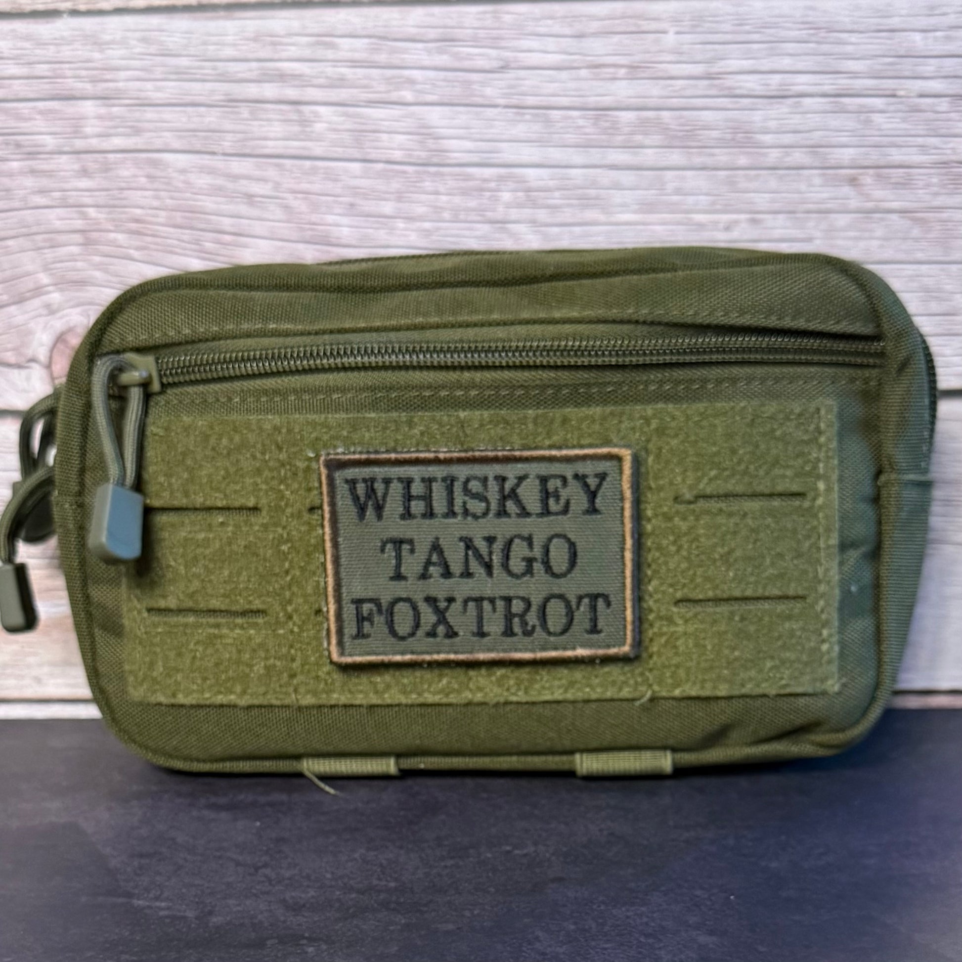 An olive green tactical pouch with a front zipper and a rectangular patch reading 'WHISKEY TANGO FOXTROT' in black letters, set against a light wood background.