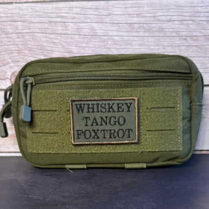 An olive green tactical pouch with a front zipper and a rectangular patch reading 'WHISKEY TANGO FOXTROT' in black letters, set against a light wood background.