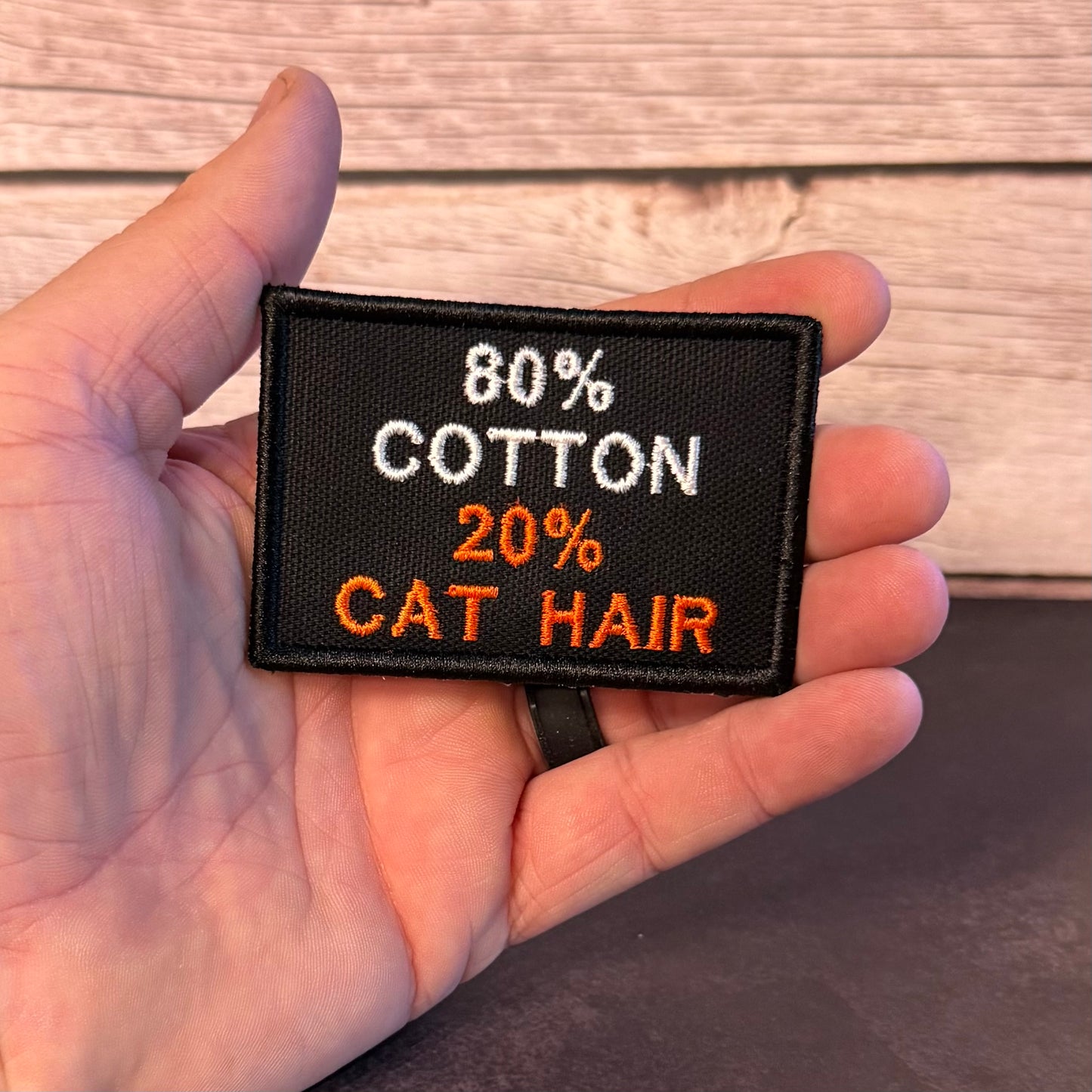 A hand holding a rectangular black embroidered patch that reads '80% COTTON 20% CAT HAIR' in white and orange text, with a wood plank background behind it.