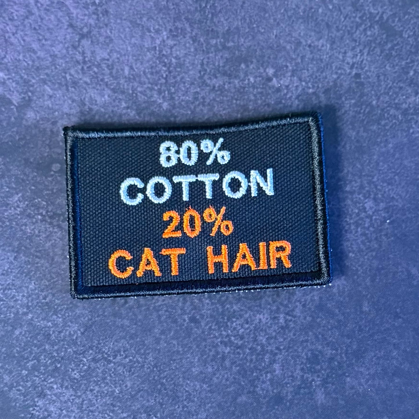 A rectangular embroidered patch with the text '80% COTTON 20% CAT HAIR' in white and orange on a dark navy background, placed on a textured blue surface.