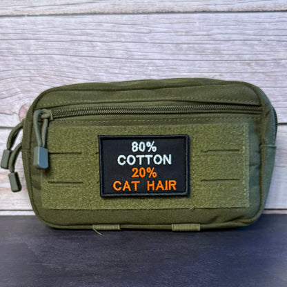 An olive green tactical pouch with a front zipper and a black rectangular patch reading '80% COTTON 20% CAT HAIR' in white and orange text, shown against a light wood plank background.