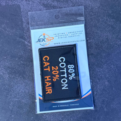 A small embroidered patch in clear retail packaging on a dark textured surface. The patch is black with orange and white text reading '80% COTTON 20% CAT HAIR'.