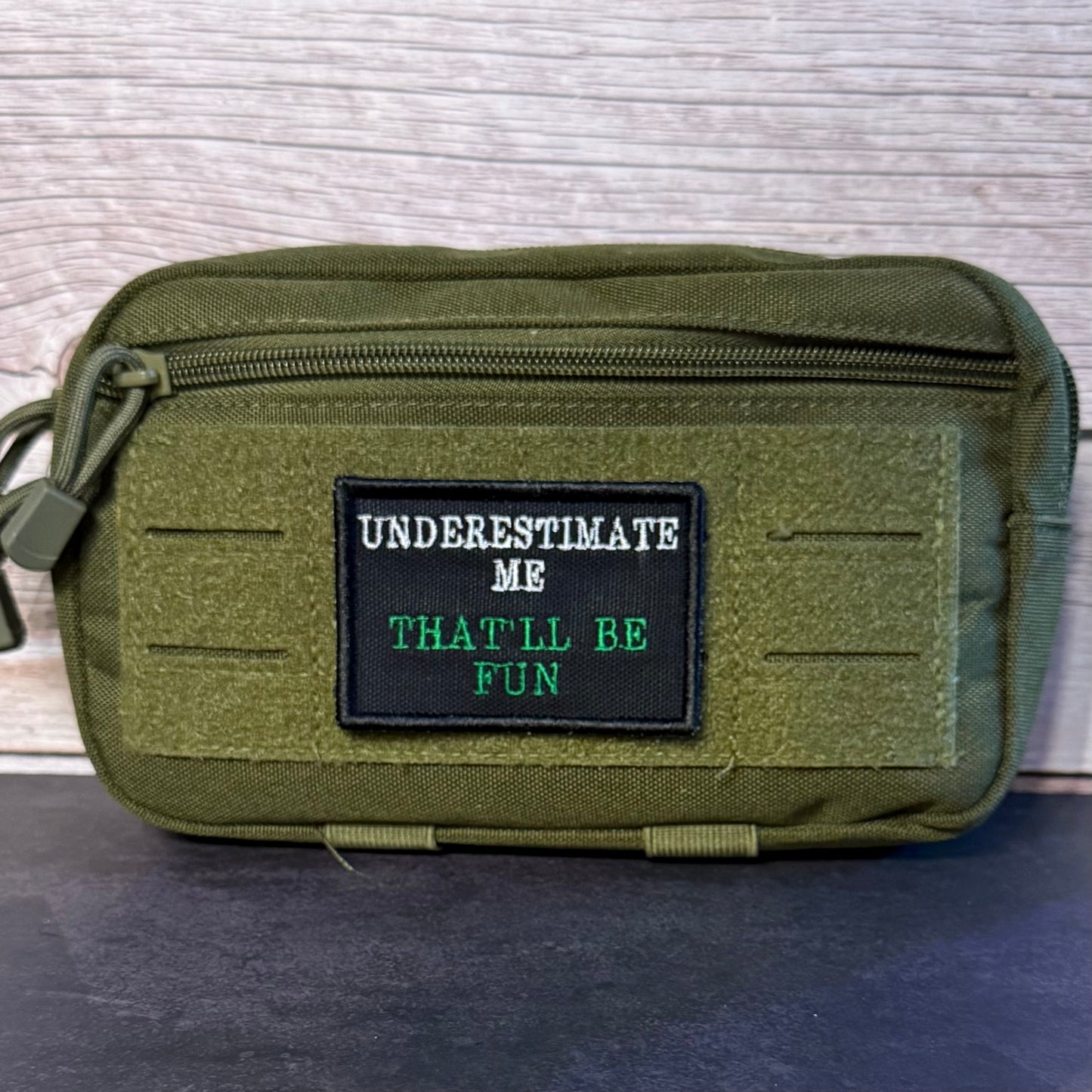An olive green tactical pouch with a black rectangular patch that reads 'UNDERESTIMATE ME THAT'LL BE FUN' in white and green text, set against a light wood background.