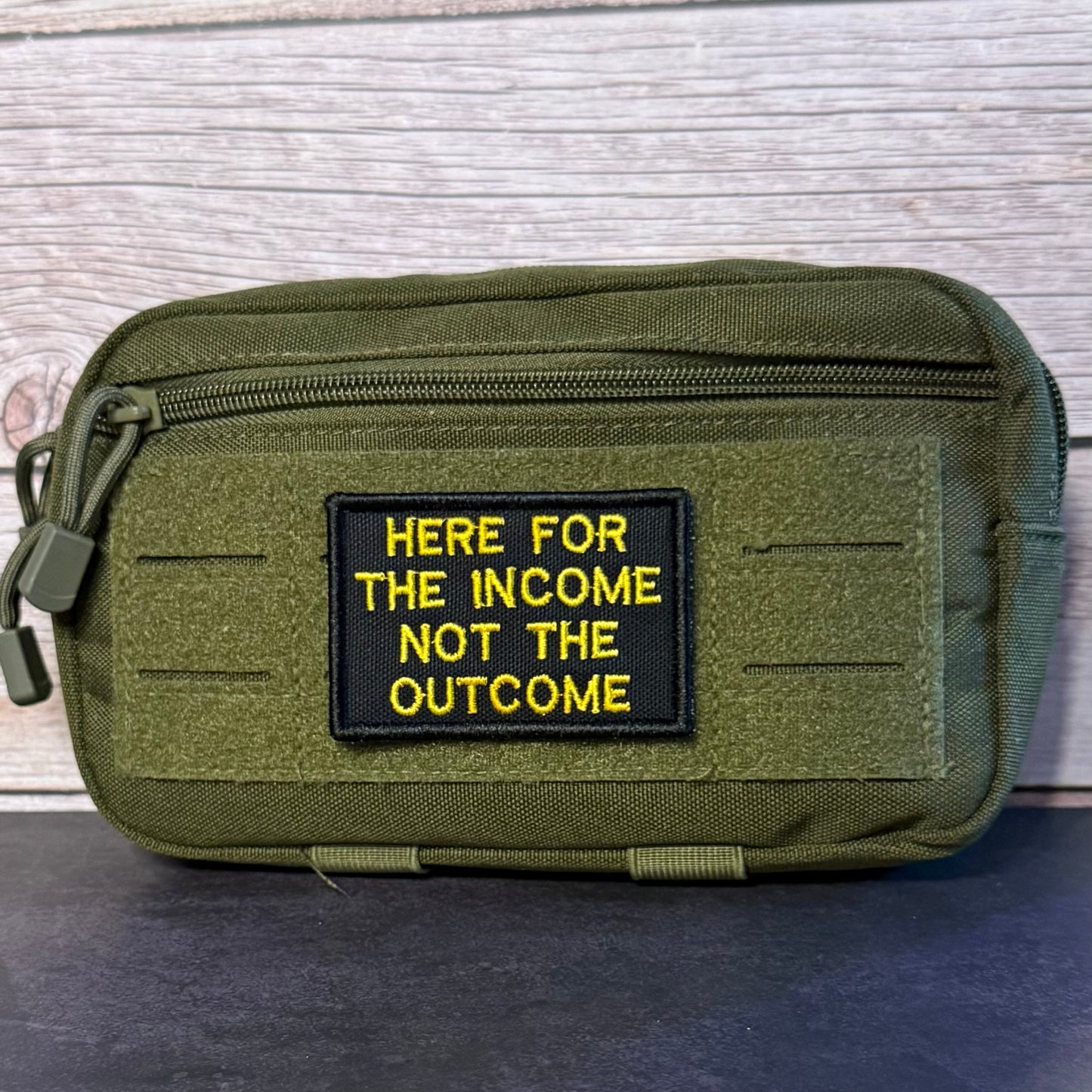 An olive green tactical pouch with a black rectangular patch featuring yellow embroidered text that reads 'HERE FOR THE INCOME NOT THE OUTCOME'. The pouch is displayed on a dark surface against a light gray wood plank background.