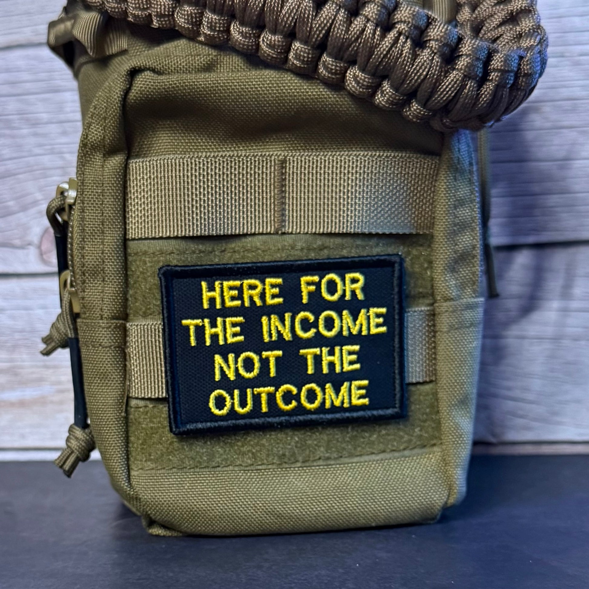An olive green tactical pouch with a black and yellow embroidered patch reading 'HERE FOR THE INCOME NOT THE OUTCOME', shown with a paracord strap against a rustic wood background.
